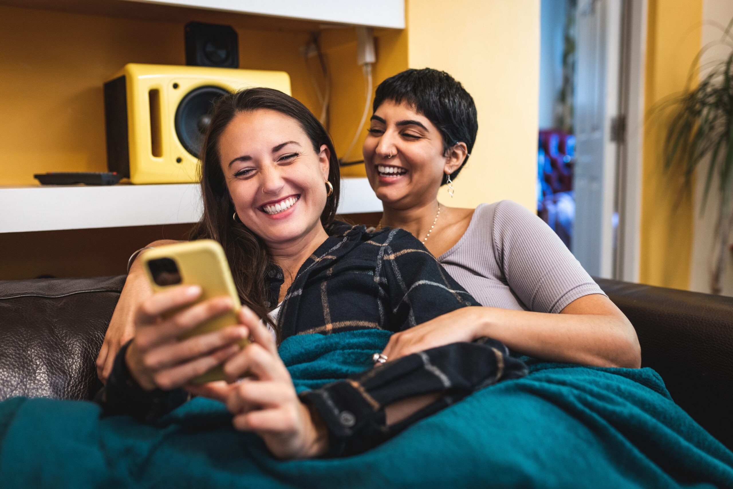 Two women cuddling on the sofa, looking at their phones and smiling